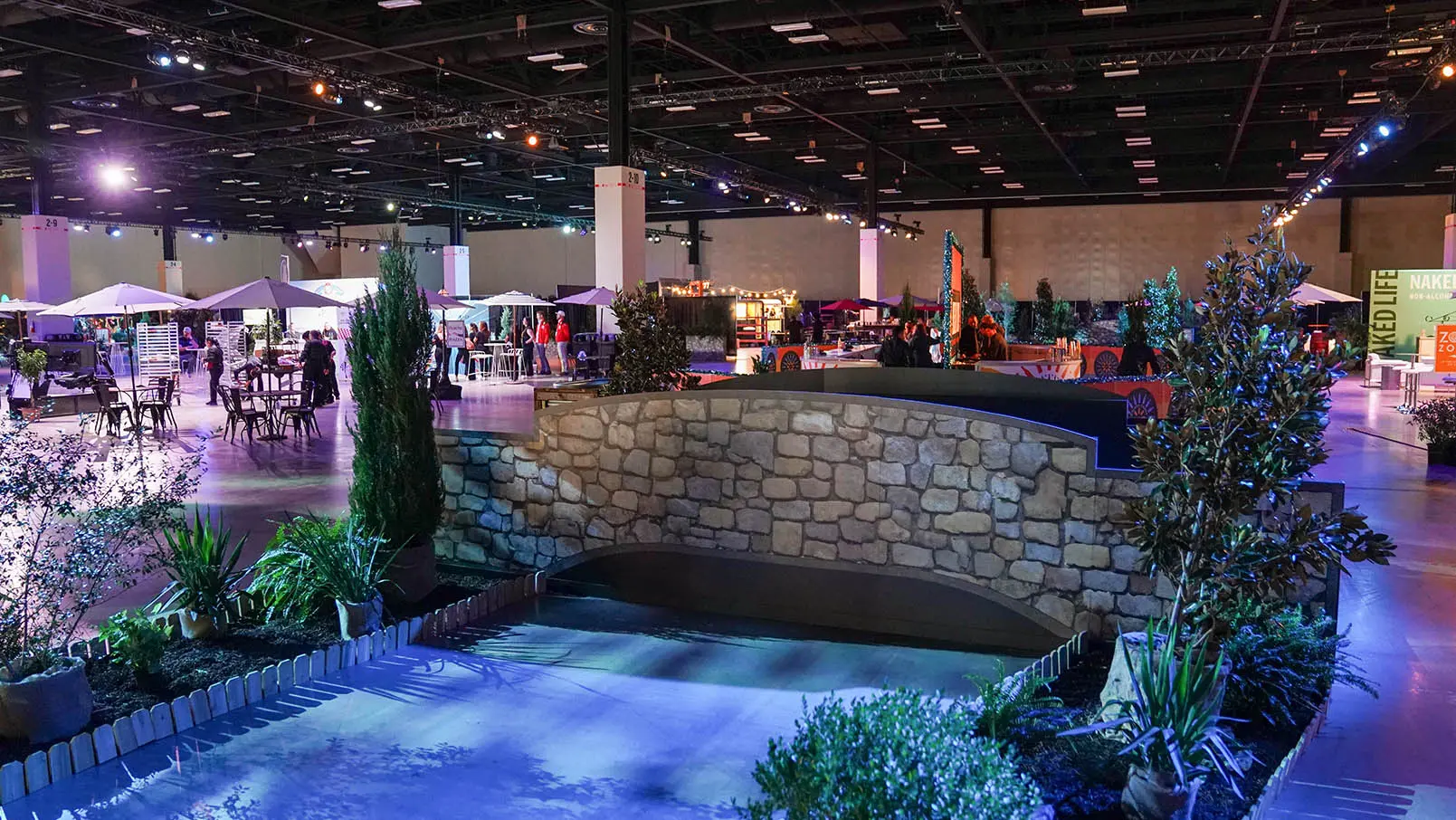 Indoor event with a replica of the San Antonio River Walk, featuring greenery and vendor booths under colorful lighting.