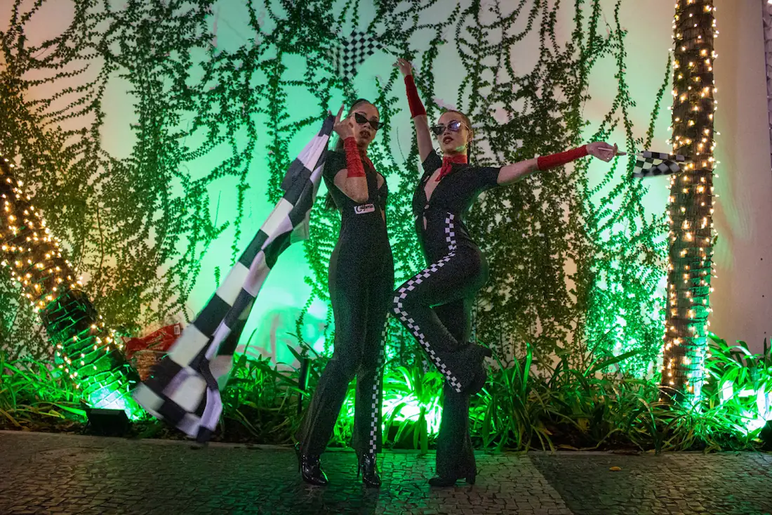 2 girls dressed in racing bodysuits carrying flags and posing in front of a decorated wall